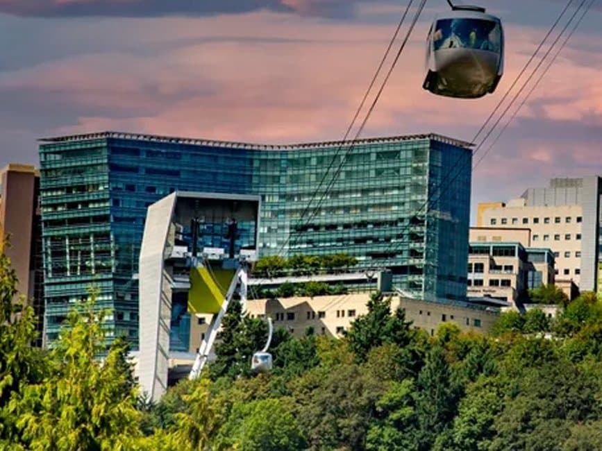 OHSU Marquam Hill campus at sunset, glass hospital buildings above the tree line with the aerial tram cabin passing overhead, Portland, Oregon